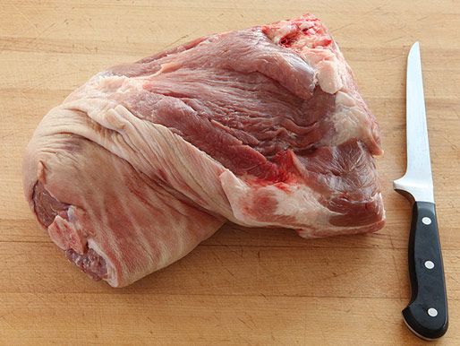 Uncooked pork shoulder on a wooden surface next to a boning knife