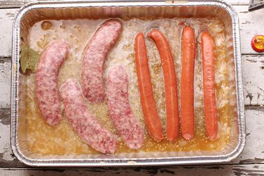 Overhead view of sausages and hot dogs partially submerged in a disposable aluminum pan filled with a flavorful braising liquid.