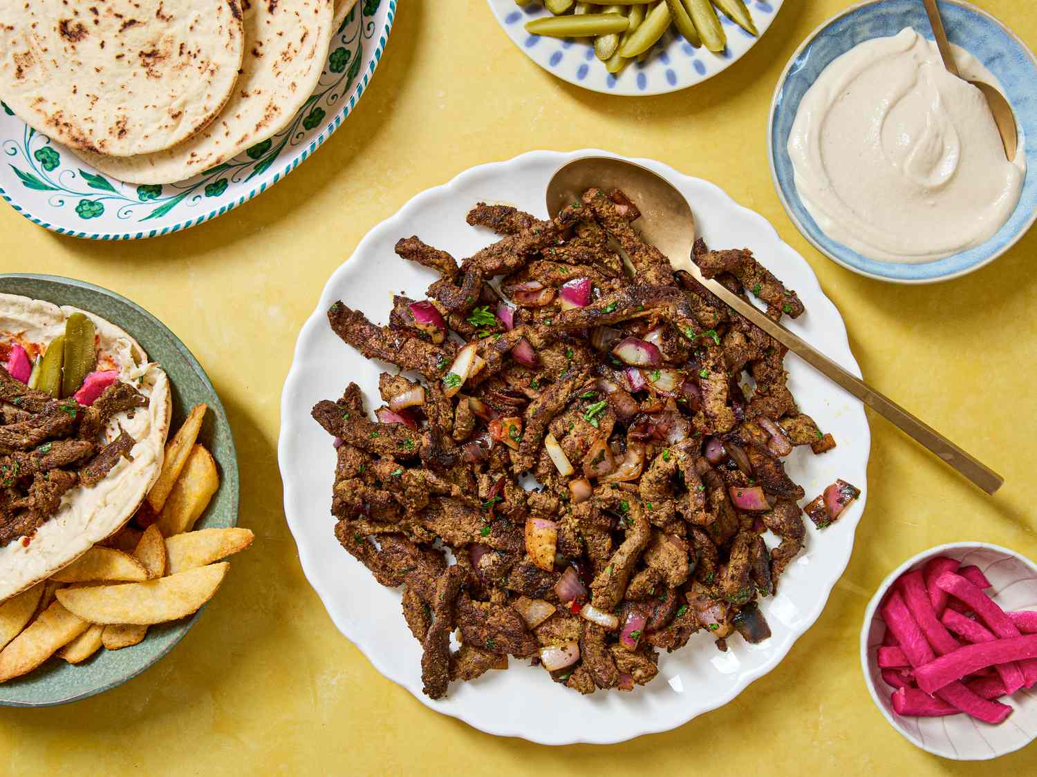 A plate of beef shawarma surrounded by accompaniments including pita fries and pickled vegetables