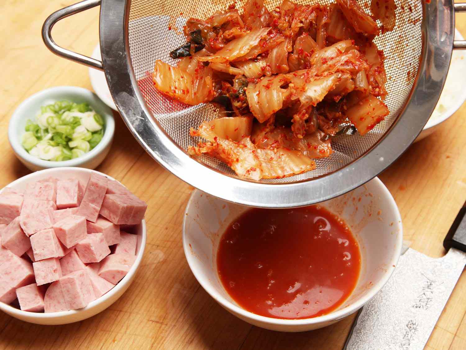 Draining out kimchi brine from a fine-mesh strainer. On the table is a bowl of cubed Spam and chopped scallions.