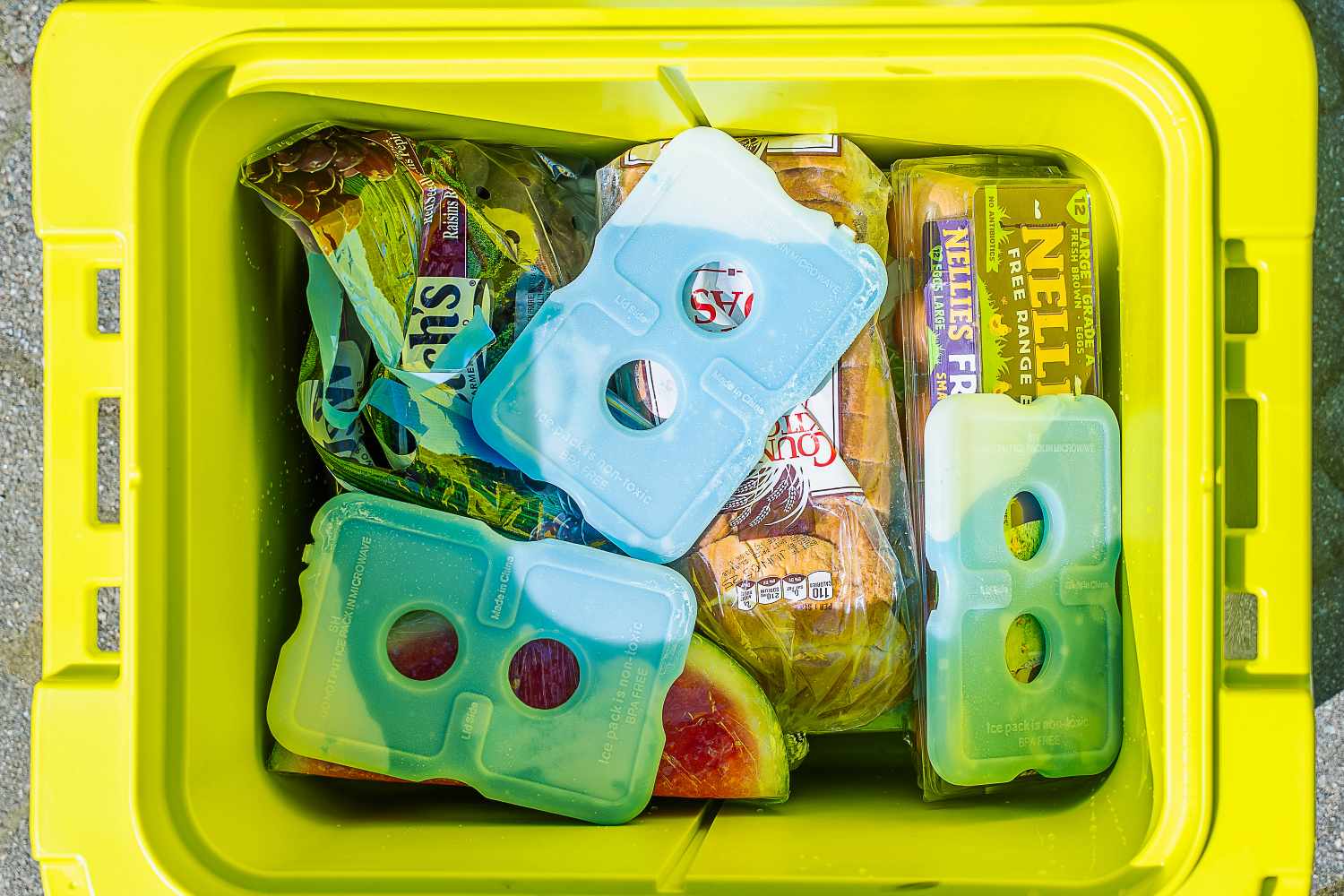 Three ice packs in a cooler with eggs, loaves of bread, and watermelon.