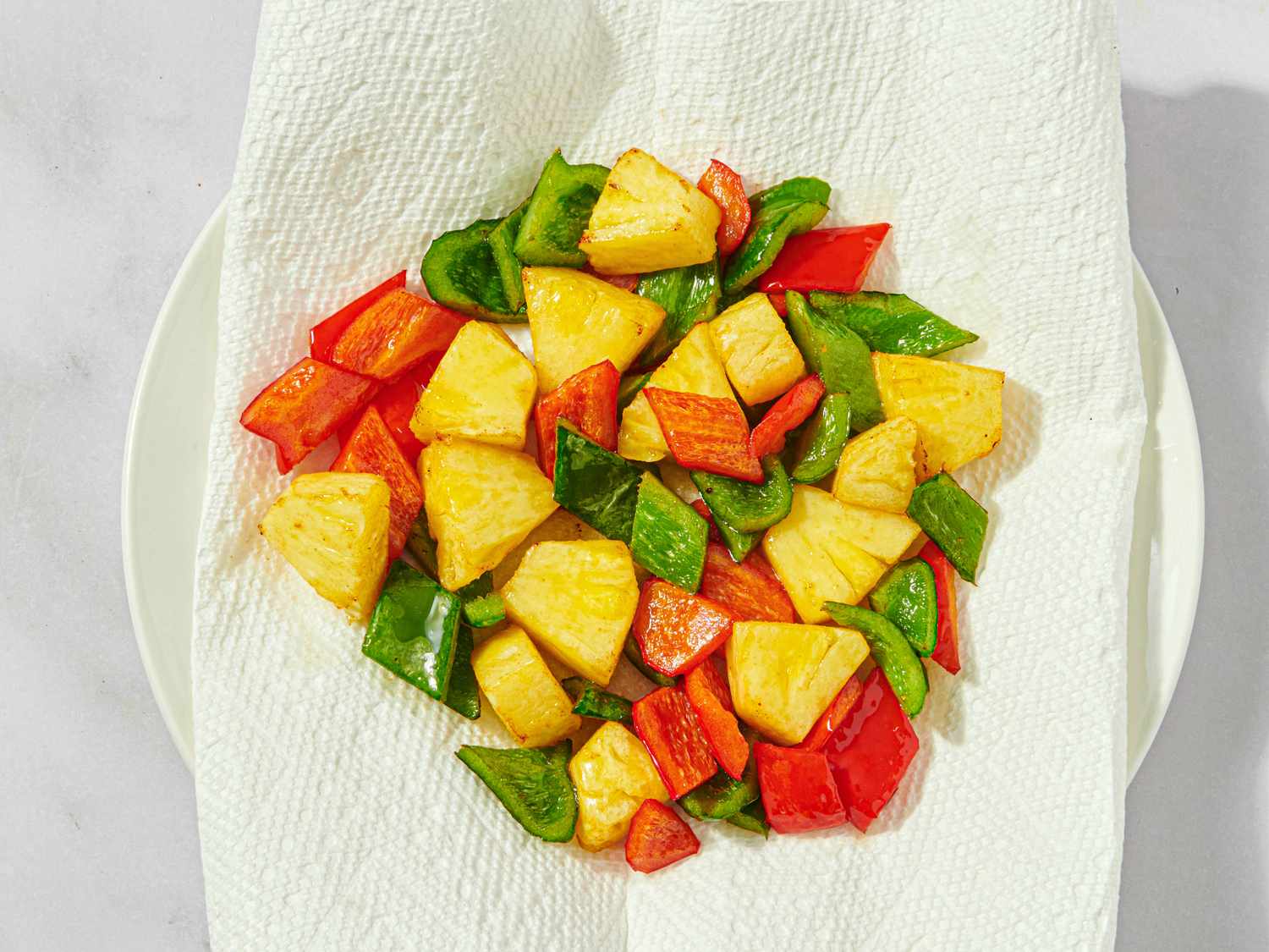 Cooked pieces of pineapple red bell pepper and green bell pepper resting on a paper towel to remove excess oil