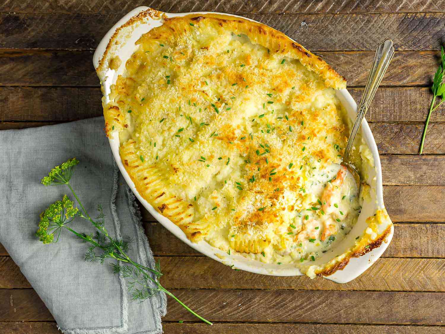 Overhead view of the fish pie served from the baking dish. A portion of the pie has been eaten, revealing the creamy sauce and chunks of fish buried beneath the mashed potato crust.