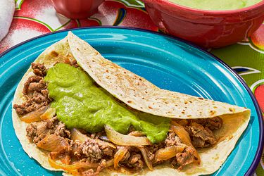 taco with mean topped with guacamole, with bowl of guacamole. Plate of tortillas, and 2 cocktails, and small dishes of onions and limes, on colorful floral printed tablecloth