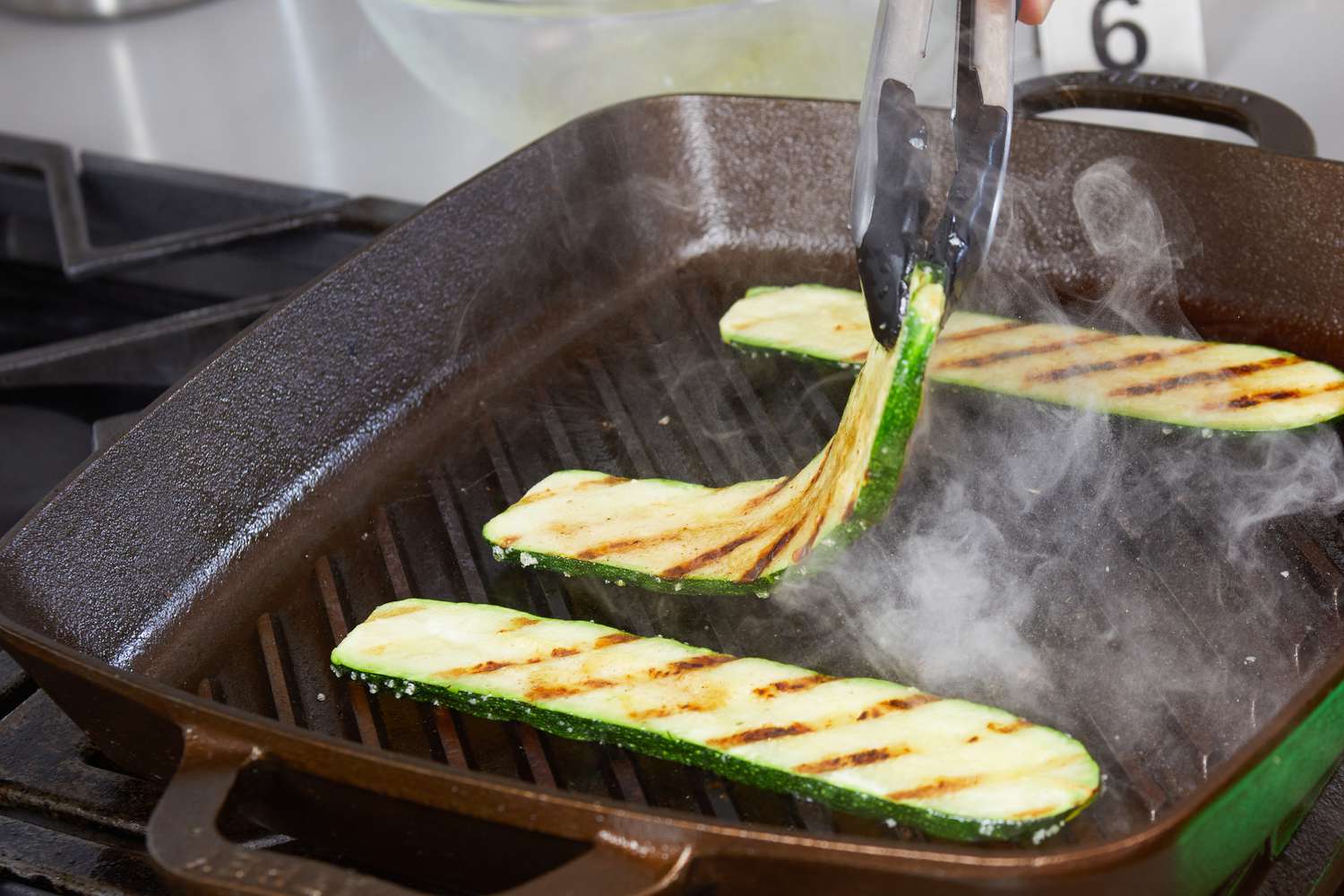 A person uses tongs to flip zucchinis being grilled using the Smithey Ironware Co. No. 12 Grill Pan.