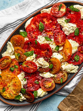 An oval ceramic platter holding caprese salad. The platter is on a dish cloth. There is a pair of forks to the left of the platter, and in the bottom right of the image is a wooden cutting board holding torn chunks of bread.
