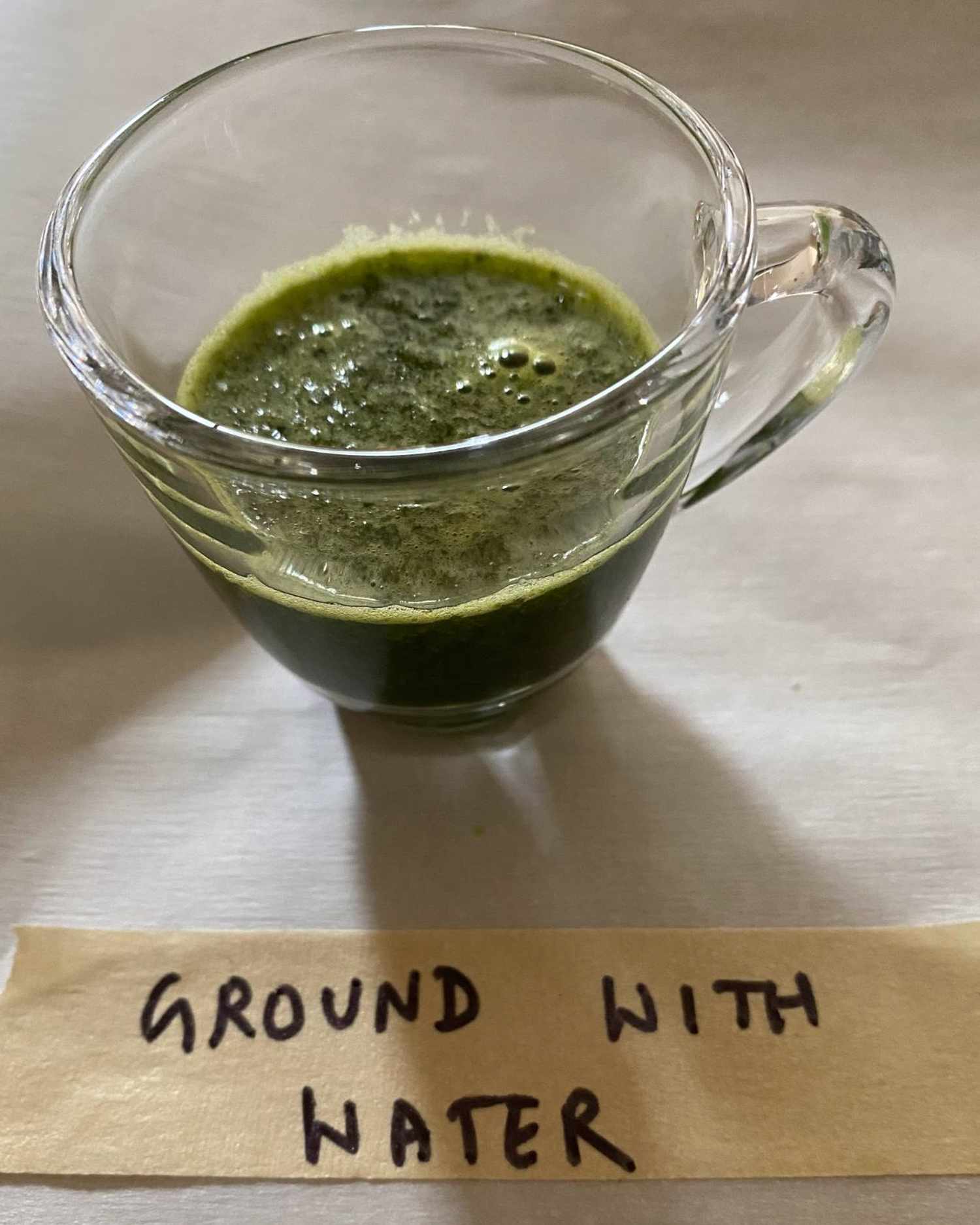 A clear cup containing a green liquid labeled Ground with Water on a handwritten tag