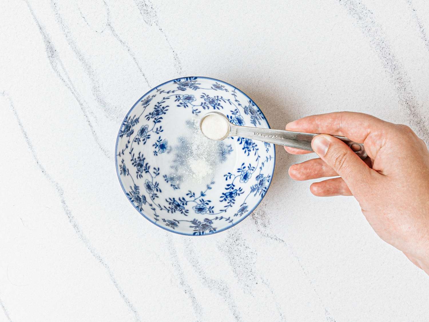 A hand placing a spoon of powdered ingredient into a patterned ceramic bowl