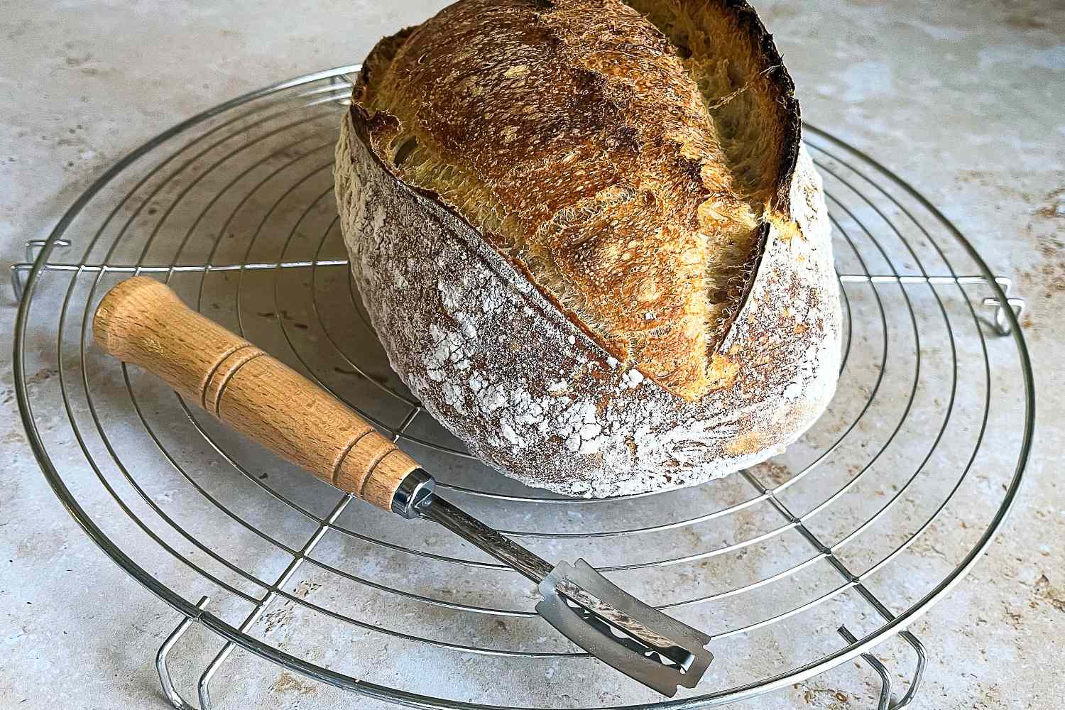 A freshly baked loaf of bread on a cooling rack with a bread lame next to it