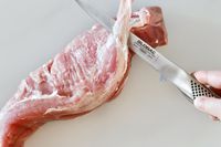A person using the Global boning knife to trim the fat off of a piece of meat.