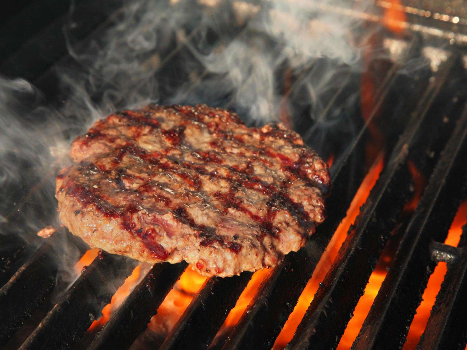 A burger patty cooking on a grill. The side facing up has cross-hatched grill marks and looks well-browned.