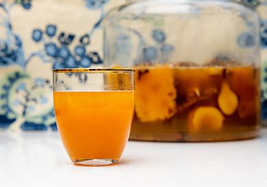 Side view of a glass of tepache next to a large batch in a gallon containter