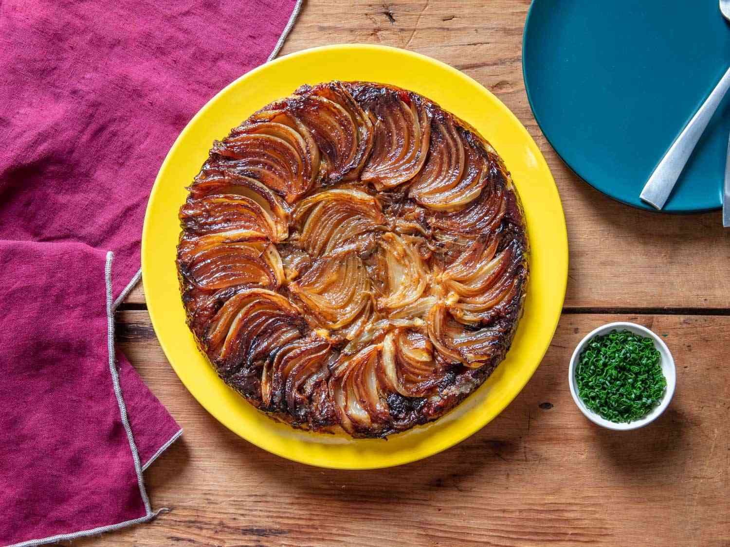 Overhead of finished tarte tatin on a yellow serving plate with a ramekin of chives next to it and a small blue plate.