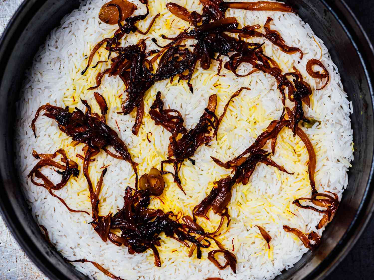 Overhead view of a pot of biryani with caramelized onions on top as garnish.