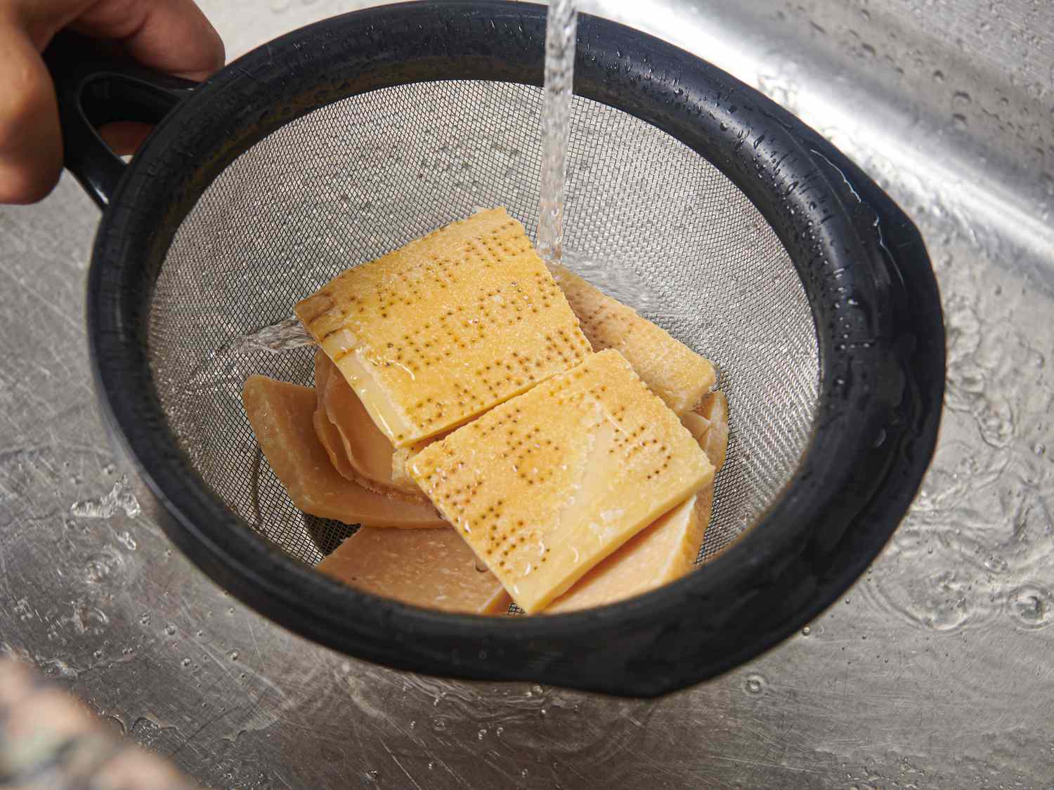 Parmesan rinds placed in a fine-mesh strainer and rinsing under water