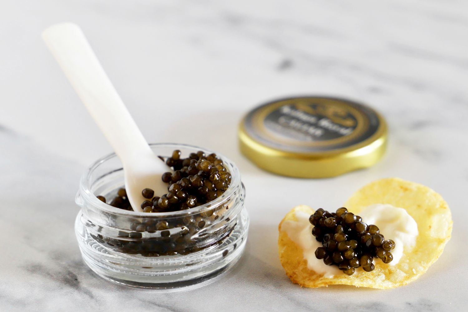 An open jar of Olma kaluga royal caviar next to a potato chip topped with crème fraîche and a scoop of caviar
