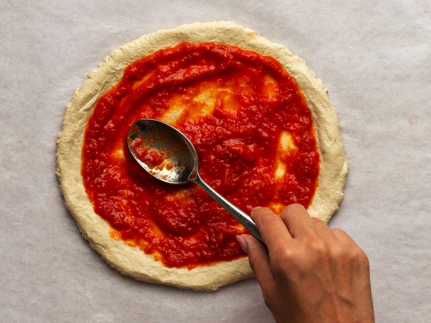 New York-style pizza sauce being spread on an uncooked pizza dough round with a metal spoon.