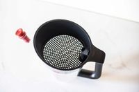 A kitchen utensil a strainer cup with a perforated lid and handle resting on a counter alongside a red stopper