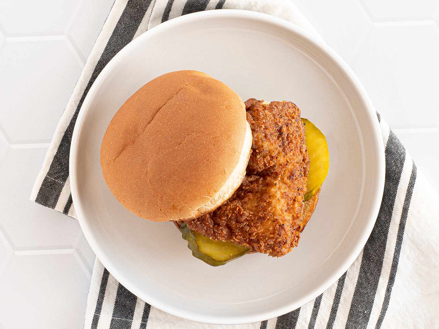 A fried chicken breast on a bun, with two slices of pickles, on a plate above a dish cloth.