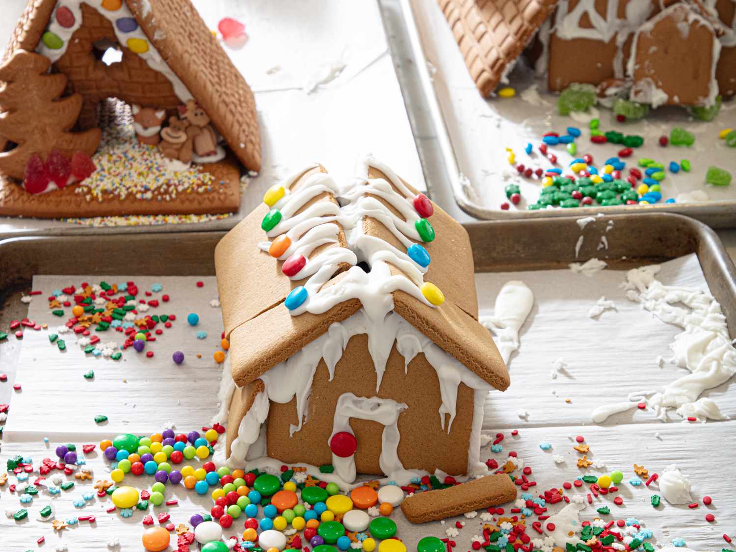 Decorated gingerbread houses with candy and icing on baking sheets and a table surface