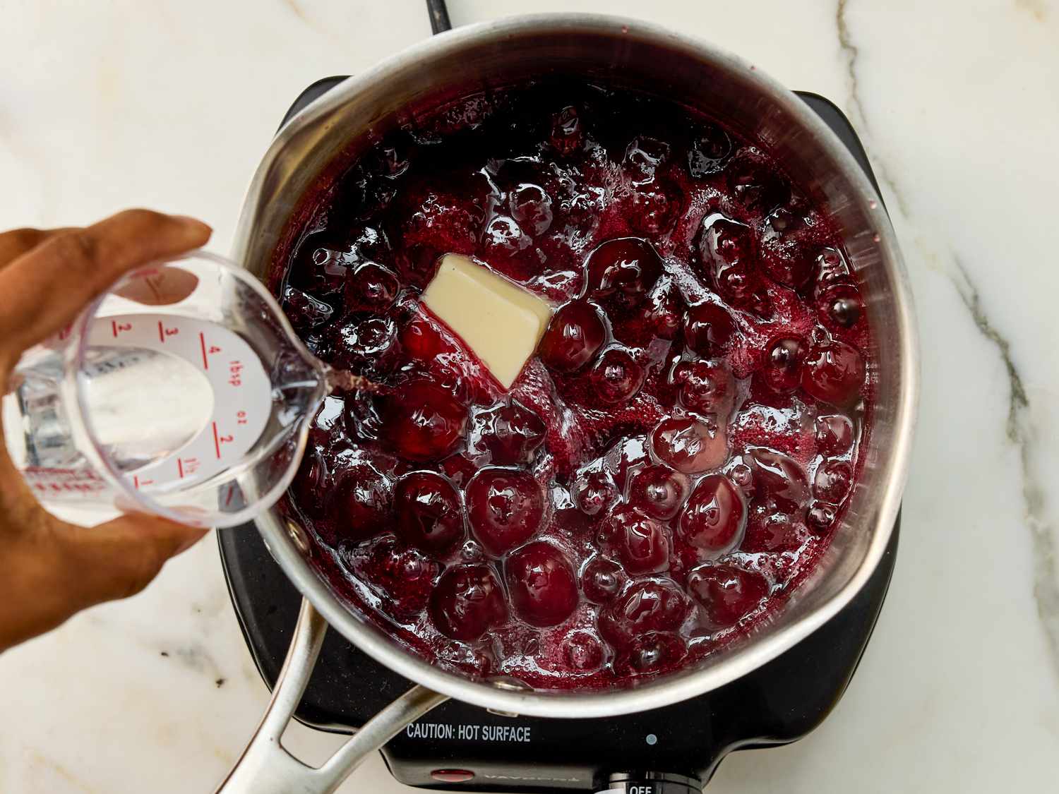 A pot on a stovetop containing cherry mixture a persons hand pouring liquid from a measuring cup into it