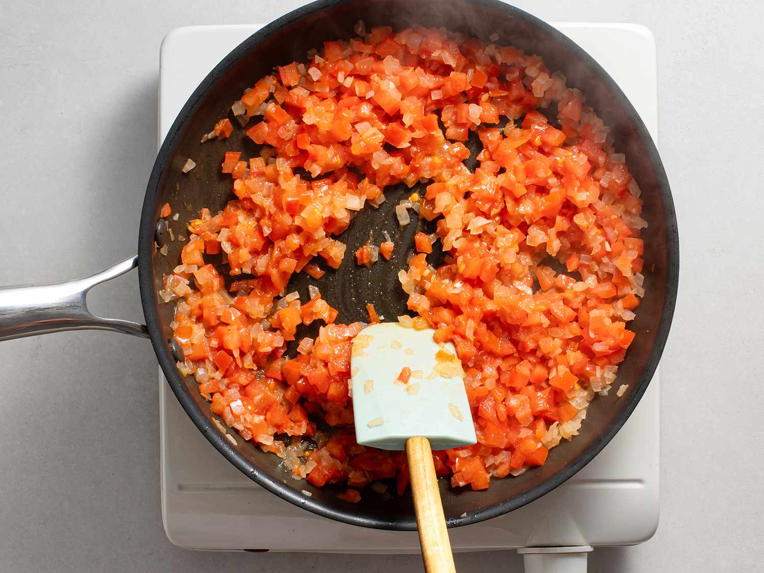 Onions and tomatoes being cooked in a non-stick pan and stirred with a silicone spatula.