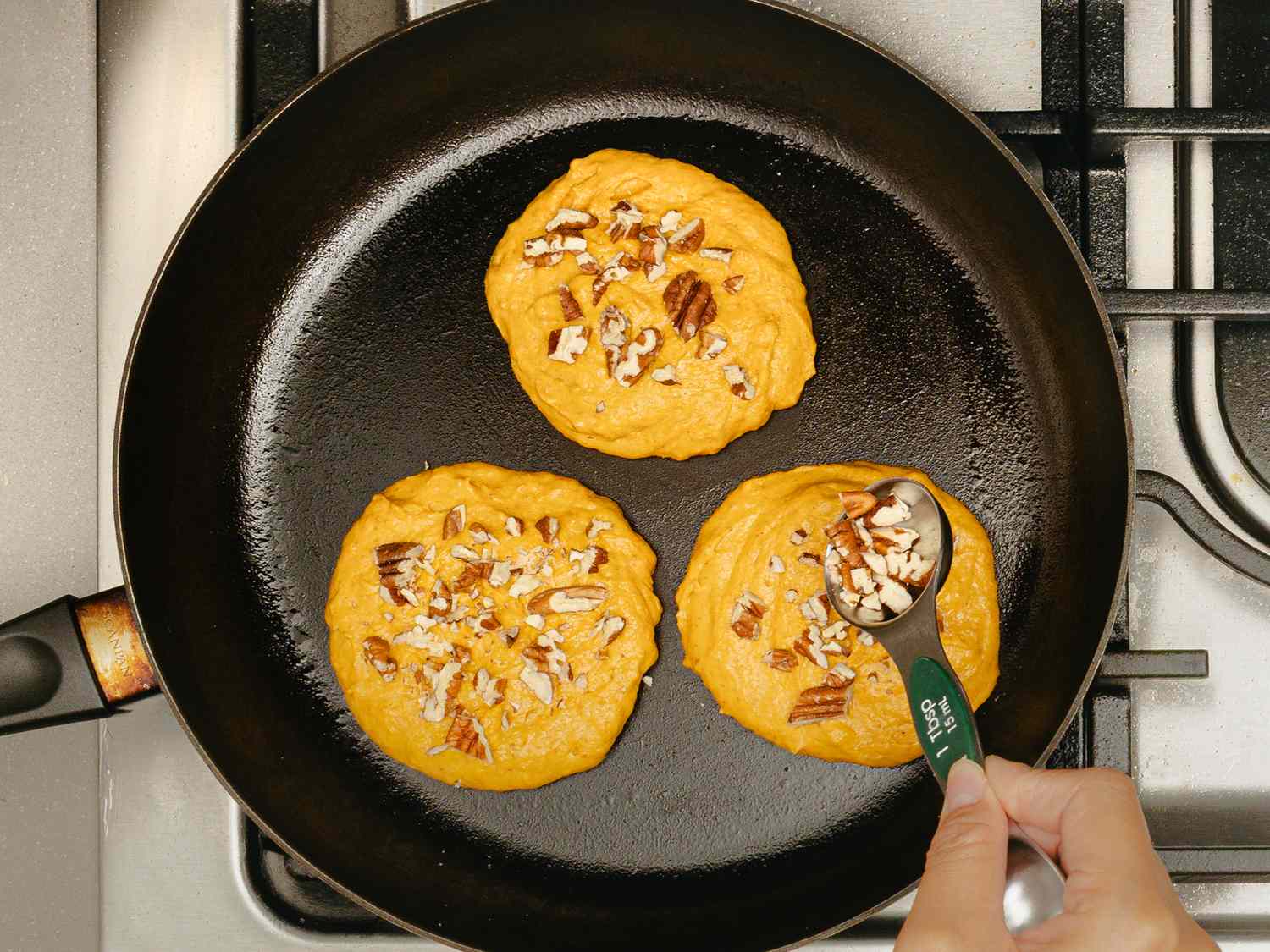 Three pumpkin pancakes cooking in a skillet with pecans a hand sprinkling nuts on one with a measuring spoon