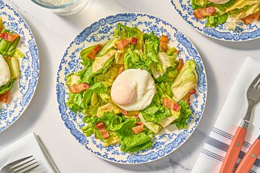 Salade Lyonnaise on antique blue and white plates with sparkling water, and utensils on white and grey striped napkin on a white marbled surface
