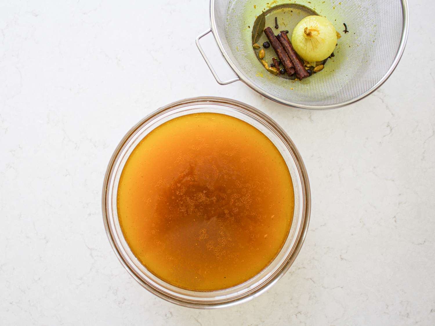 Strained broth in a glass bowl, with the strained ingredients in the strainer in the upper right of the frame