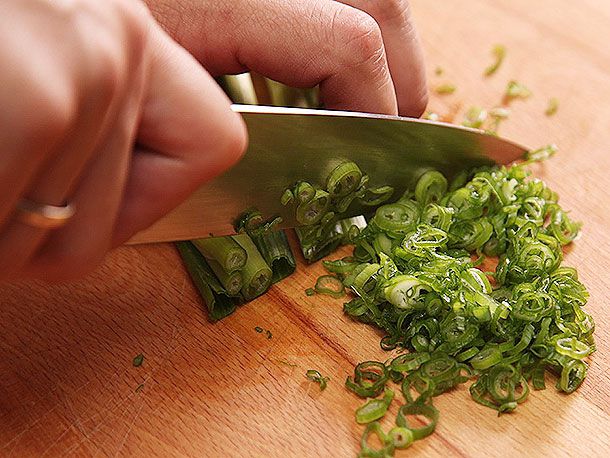 A knife held in a hand, finely slicing scallions on a cutting board.