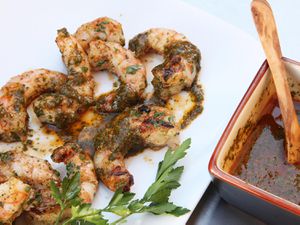 Closeup of grilled shrimp, plated on a square white platter and smothered with chermoula. A square bowl filled with more sauce is nearby.