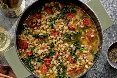 Pot of black eyed peas, kale and sausage stew, surrounded by a printed napkin, bottle and glass of wine on a stone surface 