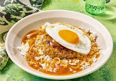 A plate of loco moco, a dish comprising rice, a hamburger patty, gravy, and a fried egg, served on a green patterned tablecloth
