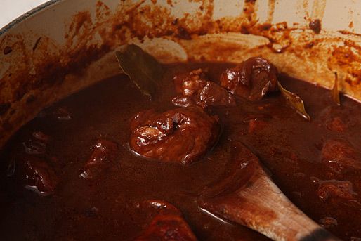 A close up of carne adovada cooking in an enamel cast iron Dutch oven. A wooden spoon is lifting up some pork and a bay leaf. 