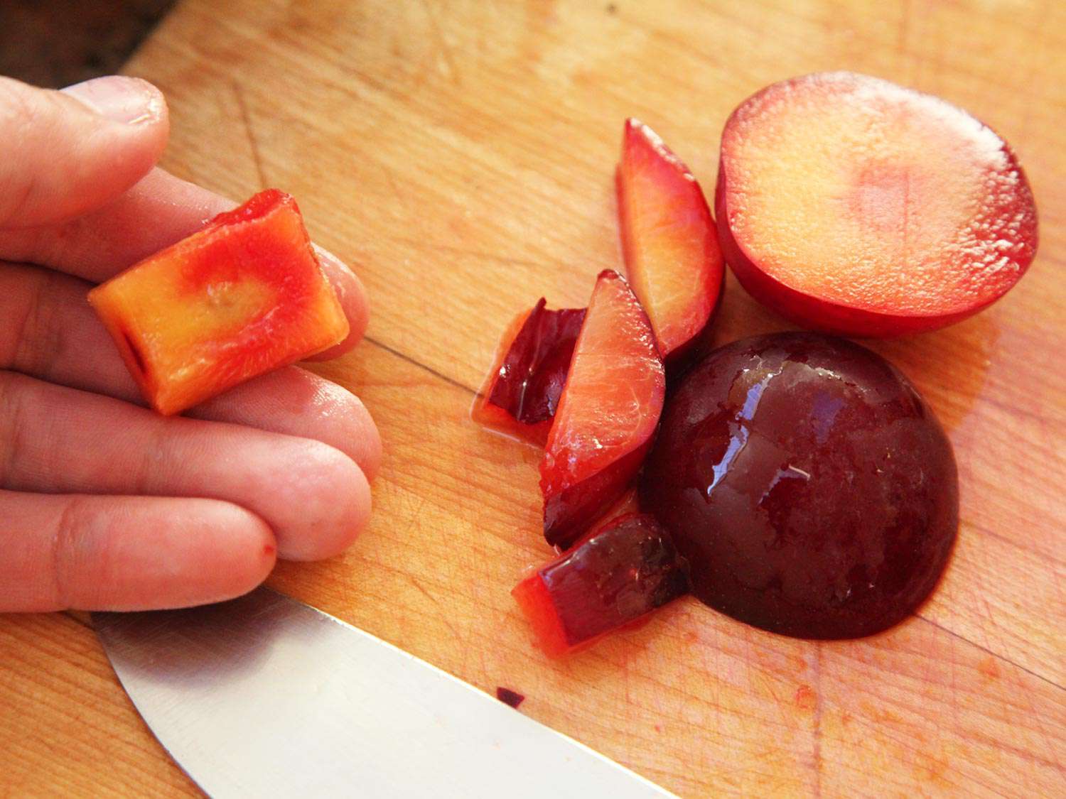 A hand holding a plum pit with some fruit surrounding it, with sliced fruit on a wooden board nearby.