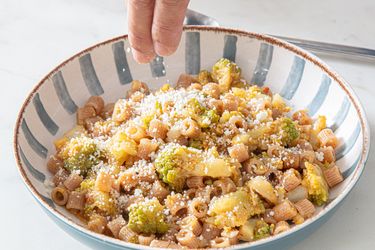 A bowl of pasta with Romanesco broccoli being sprinkled with grated cheese by a hand