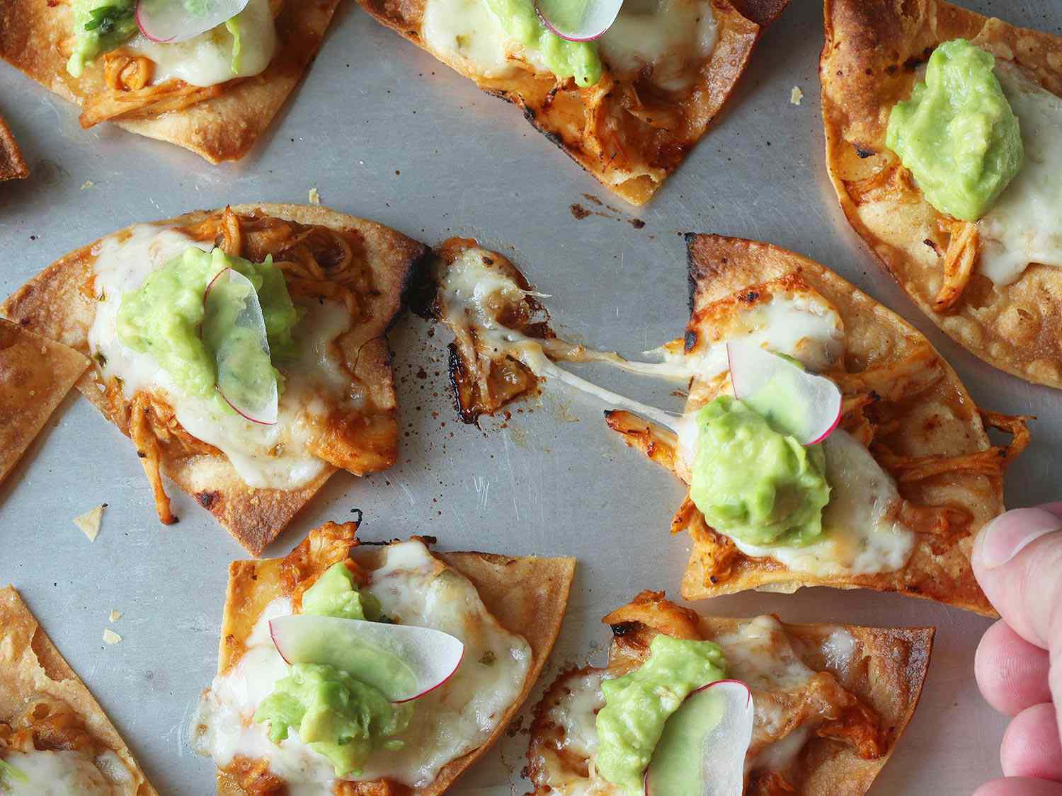 Pulling a nacho from a sheet pan of chicken tinga nachos topped with guacamole and sliced radish.