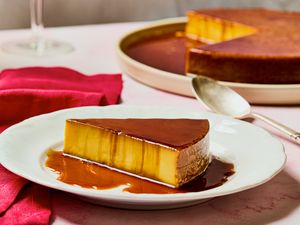 A slice of orange and saffron flan on a plate with caramel sauce placed beside a full flan serving in the background