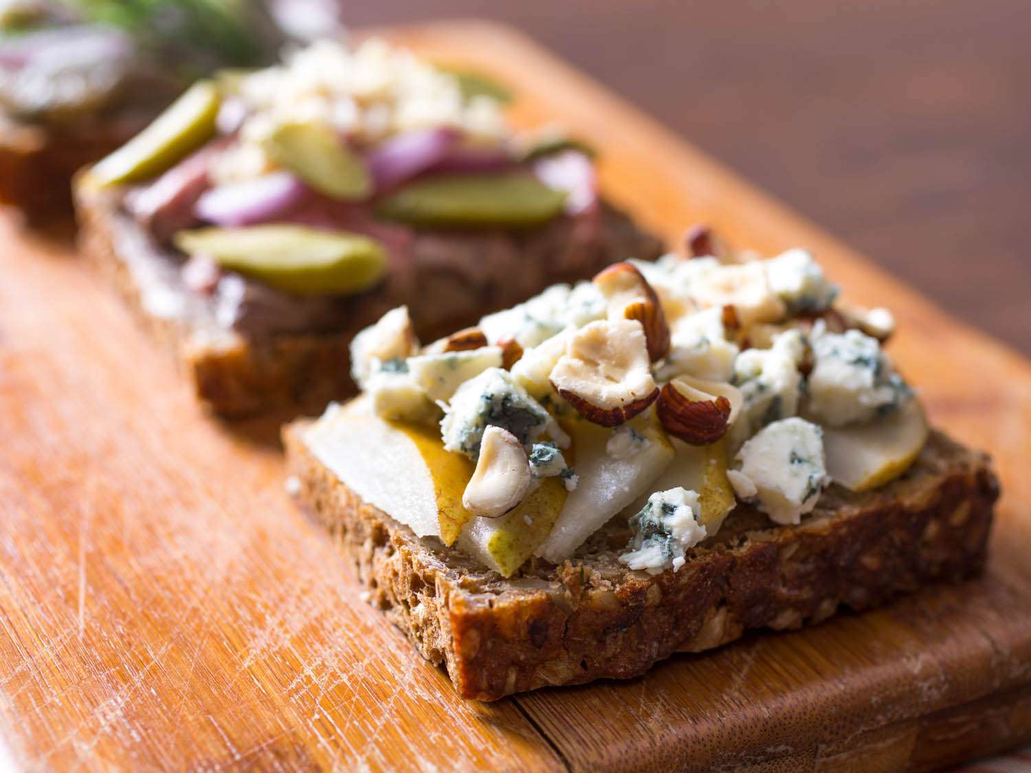 Three different Smørrebrød on a wood cutting board, each with different toppings. A Smørrebrød with pears, hazelnuts, and blue cheese is in the foreground. 
