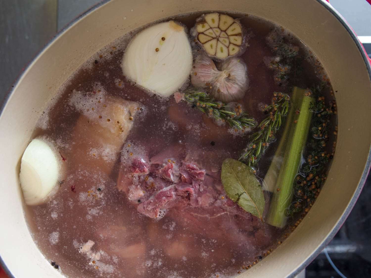 overhead view of meats, clove-studded onion halves, coriander seed, peppercorns, bay leaf, rosemary, celery, and halved garlic head in Dutch oven