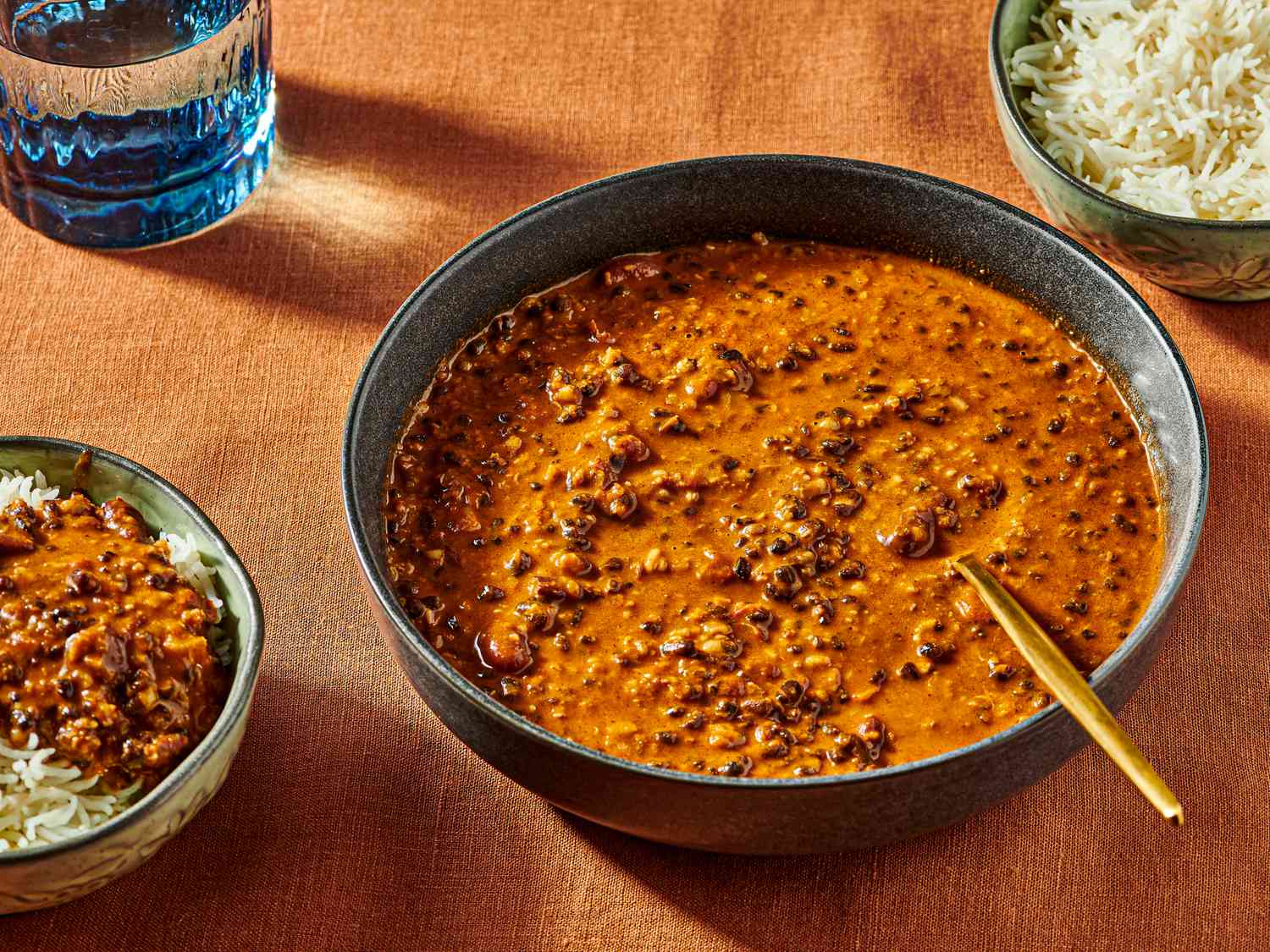Dal Makhani in a bowl with a spoon, with rice in the top corner, and a smaller bowl of dal and rice. Burnt orange colored surface and blue glass of water. 