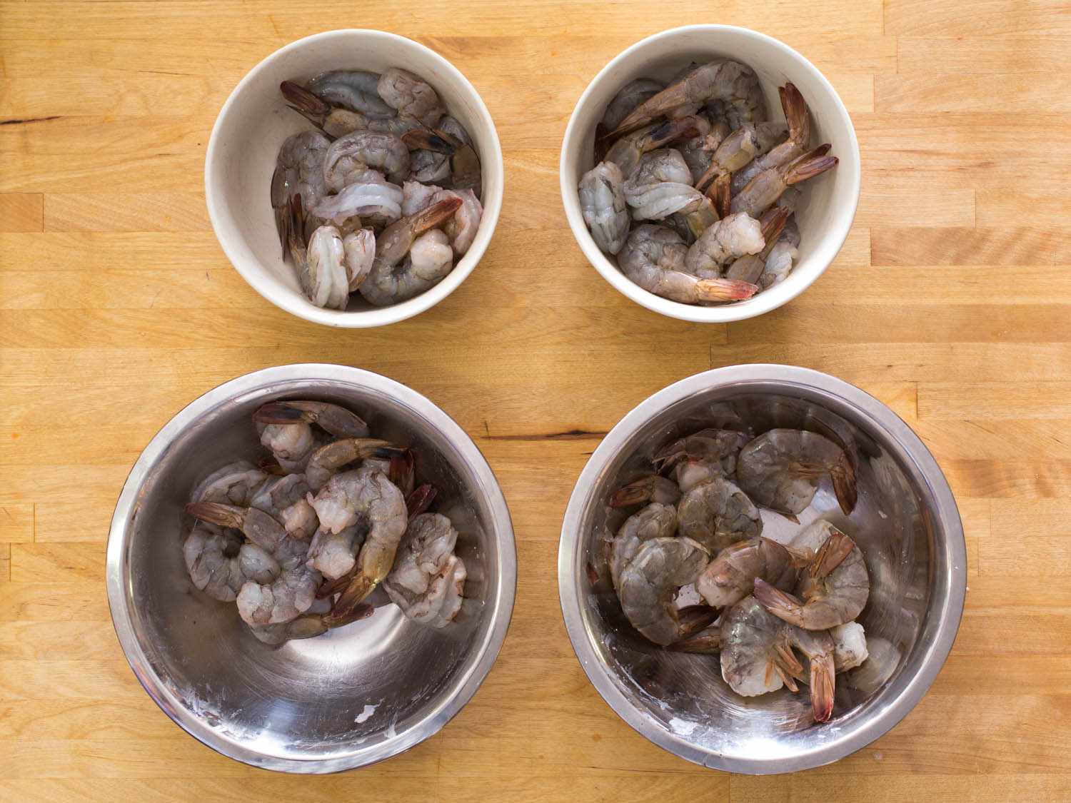 Two silver bowls and two smaller white bowls of raw shrimp resting on a light wooden countertop.