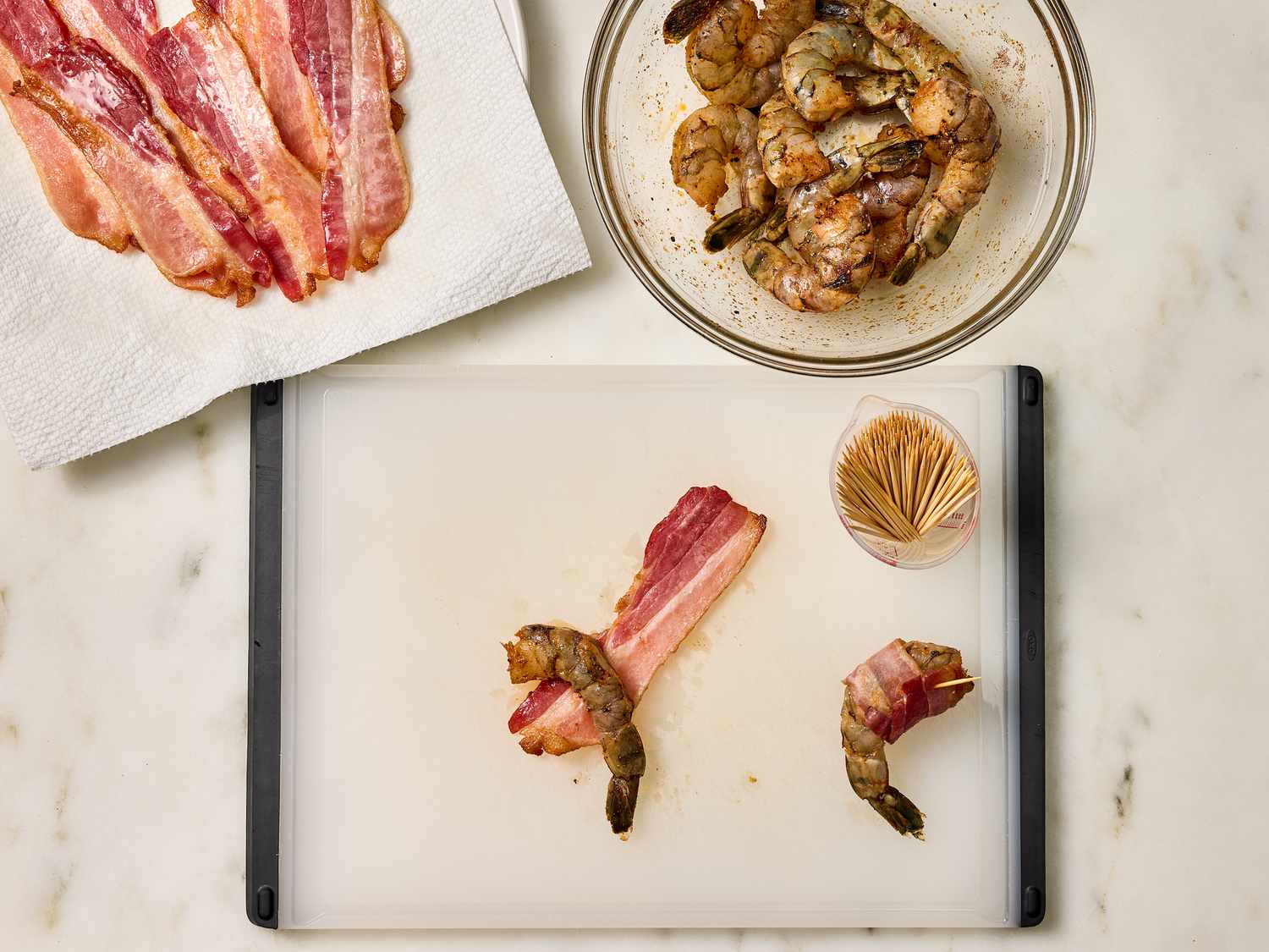 Shrimp on a chopping board, being wrapped in bacon. Plate of bacon and bowl of shrimp on the side