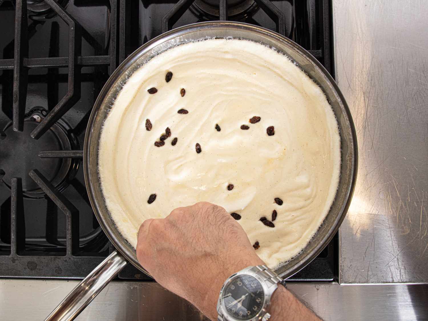A hand stirring batter with raisins in a skillet on a stove
