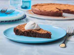 Slice of flourless chocolate cake, topped with a dollup of cream, on a blue plate, blue tabletop, and the cake is in the background 