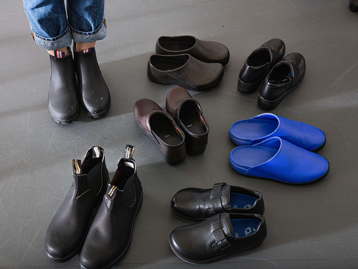 Various pairs of kitchen shoes. A person wearing boots standing beside the shoes.