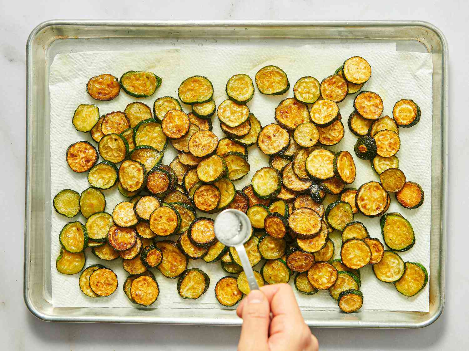 A tray of roasted zucchini slices being sprinkled with salt from a spoon