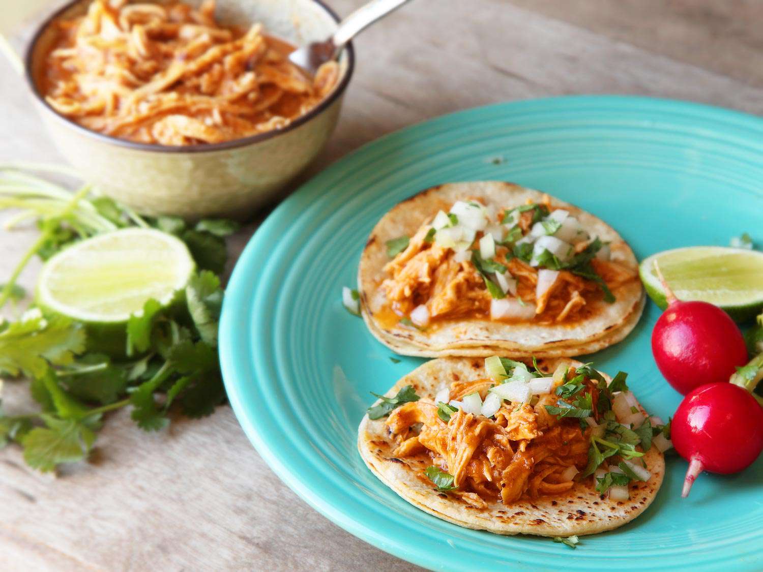 Two tacos filled with chicken tinga on a plate with some radishes and lime wedges. 