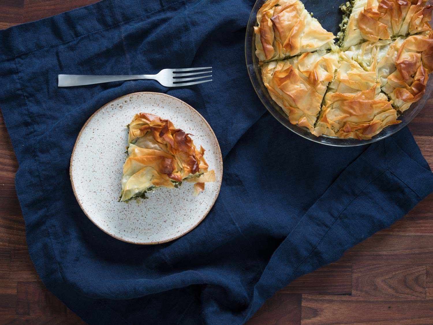 A wedge of spanakopita on a plate, next to a pie dish of spanakopita