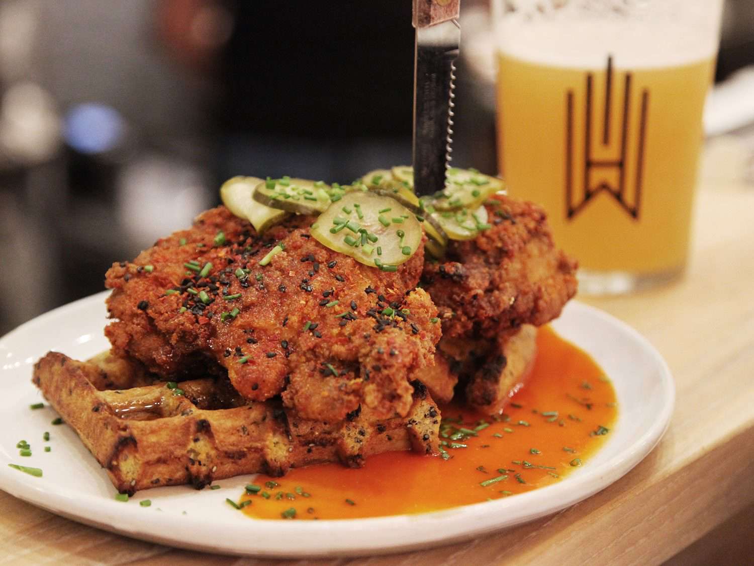 Kimchi-brined fried chicken, topped with dill pickle slices, atop a scallion cornmeal waffle, on a white plate, with a knife stuck vertically in the chicken breast and a glass of beer in the background.